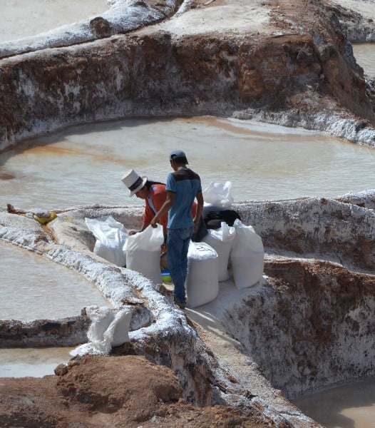 two workers, one in traditional clothing working on the Maras Salt Pans, picking up 70kg sacks of salt to carry up to the storage room.