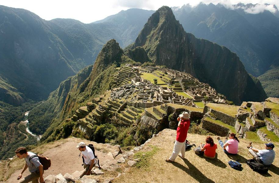An aerial view of Machu Picchu reveals the ancient citadel nestled amidst lush greenery and towering peaks. Tourists dot the terraces and pathways, captivated by the site's beauty. The sun shines, casting a golden glow on the stone structures, while the surrounding mountains create a stunning backdrop. It's a picture-perfect day at this remarkable wonder of the world.