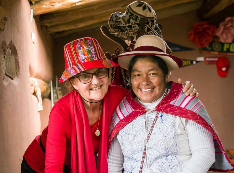 this picture is showing a peruvian woman in traditional clothing an a hat together with an old lady tourist smiling into the camera. they both are in a room together.