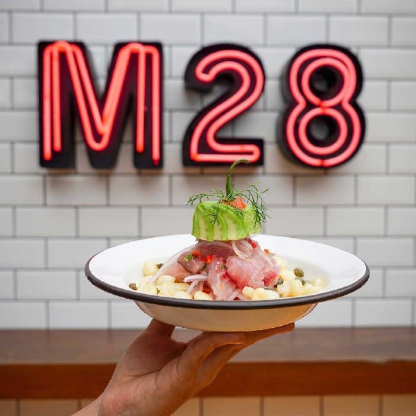 In the picture, the vibrant logo of Mercado28 (M28) catches the eye with its bold and stylish design. The logo features the letters "M28" in a modern font, creatively intertwined to form a cohesive emblem. Positioned in front of the logo is a tantalizing plate of ceviche, a popular dish in Latin America. The ceviche showcases a medley of fresh seafood, tangy citrus flavors, and colorful garnishes, enticing the taste buds. The logo and the ceviche together represent the essence of Mercado28, a culinary destination where traditional flavors and contemporary aesthetics come together to create a delightful experience for food enthusiasts.