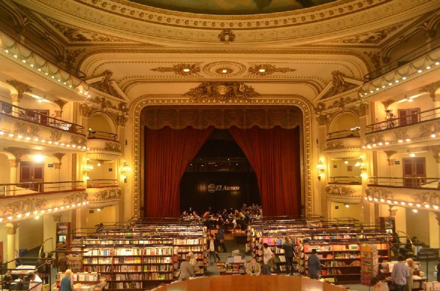 The picture showcases the grandeur of El Ateneo, a renowned bookstore in Buenos Aires, Argentina. As you step into the store, you are greeted by a breathtaking sight: a vast theater room that has been transformed into a literary haven. The room exudes an air of elegance and nostalgia, with its ornate architectural details and opulent golden lighting that casts a warm glow throughout the space. In the center of the room, a vibrant red curtain hangs, creating a focal point that adds to the dramatic ambiance. The rows of bookshelves, filled with countless volumes, line the walls, inviting visitors to explore the vast literary treasures within. The combination of the theater-like setting, the golden light, and the majestic red curtain creates a captivating and enchanting atmosphere, making El Ateneo a truly unique and awe-inspiring destination for book lovers and cultural enthusiasts alike.