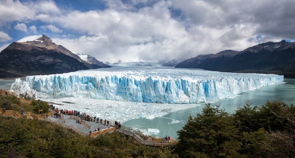 Perito Moreno Glacier in Argentina's Los Glaciares National Park, tourists standing on the wooden viewing platform overslooking the massive glacier that spans across the entire image from left to right