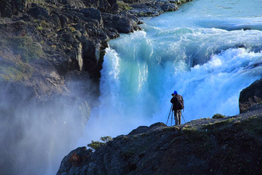 The picture depicts a mesmerizing scene at the edge of a cliff near the majestic Salto Grande waterfall in Torres del Paine National Park, Chile. A lone tourist stands captivated, gazing at the powerful cascade of water tumbling down the Paine River. The breathtaking landscape is adorned with lush natural vegetation, showcasing the diversity of the surrounding flora. In this tranquil setting, one may also catch a glimpse of the wild guanaco, adding to the allure of this remarkable destination.