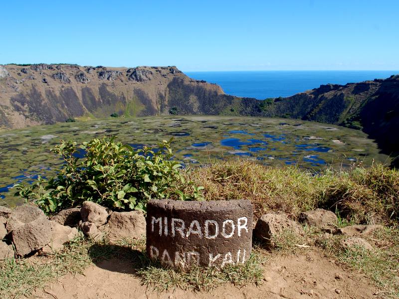 Volcano crater lookout point with a sign featuring its name 'Rano Kau'. The crater is filled with water and algae overlooking the Pacific Ocean 