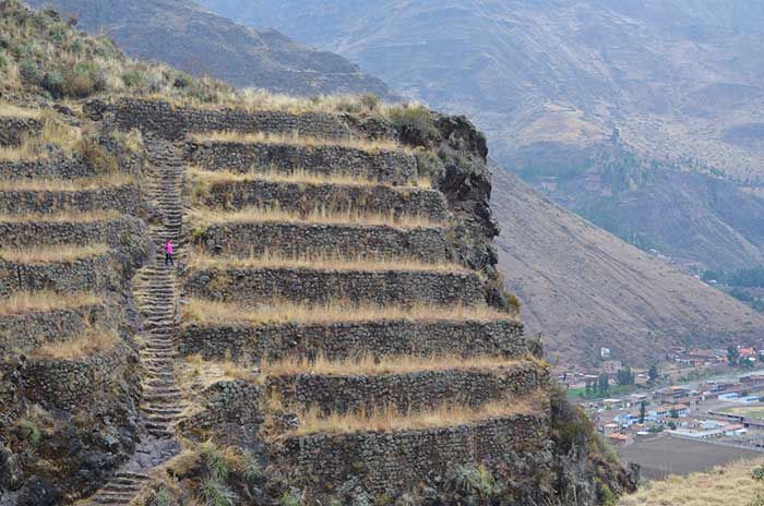 A tourist wearing a pink jacket hiking up to the top at Pisaq Ruins with Pisaq town as backdrop (Sacred Valley Cusco) 