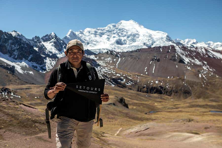 Man holding a Setours flag and the Ausangate snow-capped mountain as backdrop. Along the trail to Vinicunca Rainbow Mountain