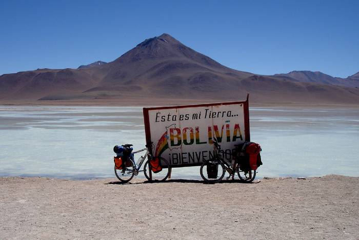 A welcome board on the Chilean Bolivian border with 2 backpacker bikes and a mountain and the white lagoon as backdrop  