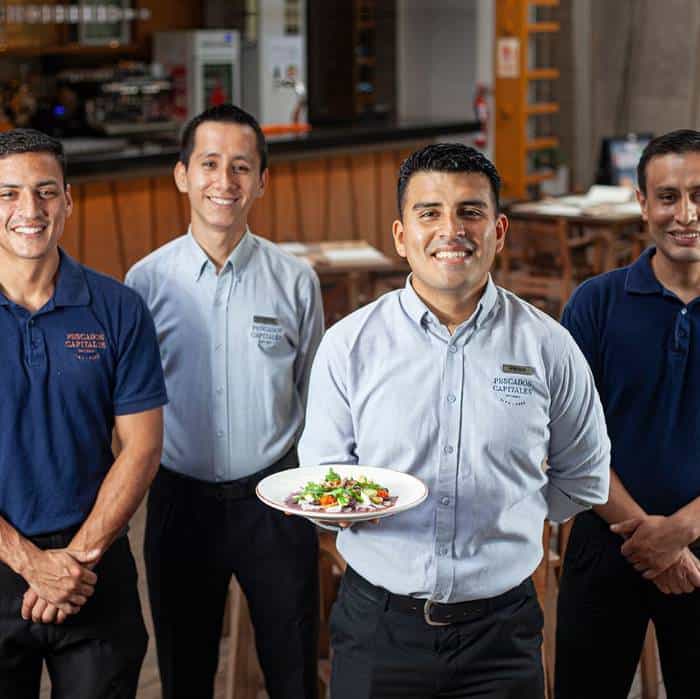 Four male waiters smilinh and one of them showing a plate featuring a Peruvian ceviche at the cevicheria Pescados Capitales