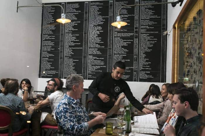a waiter showing the restaurant's menu to guests sitting at a table and a huge board featuring an inmense list of wine by the glass at BocaNariz, Santiago de Chile