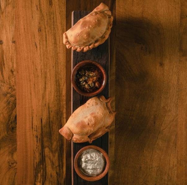 Two empanada filled pastries served on a wooden table with 'chimichurri' sauce and a white herbed cream sauce  