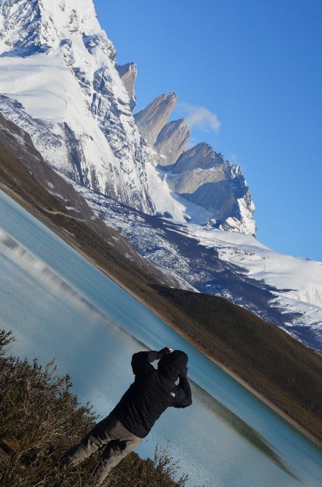 photographer standing next to lake in a black jacket and taking a picture of the Paine Towers behind snow-capped mountains with a blue sky in the background