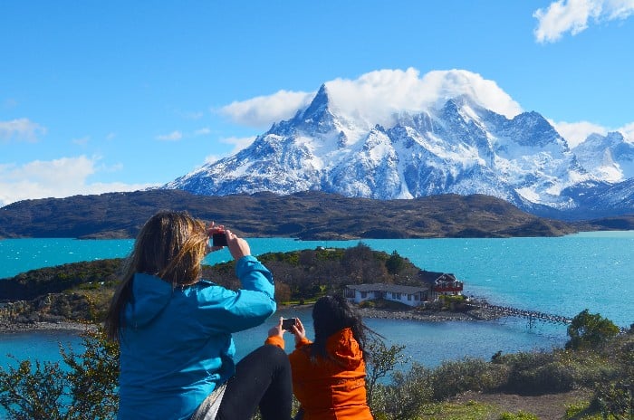 Two people taking pictures atop a hill overlooking Pehoe Lake with snow covered mountains in the background in Chilean Patagonia Torres del Paine National Park