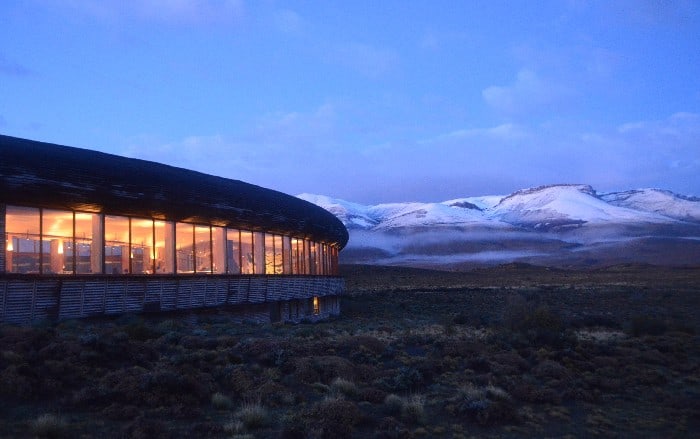 Facade of Tierra Patagonia on the left-hand side after sunset with the internal lights turned on with orange glow and snow-capped mountains in the background 