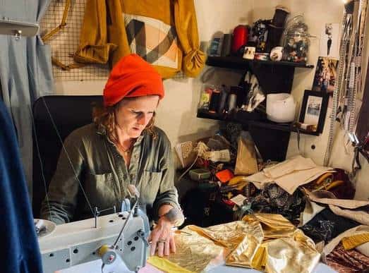 a woman sewing a piece of garment surrounded by thread rolls, other handmade clothing and  